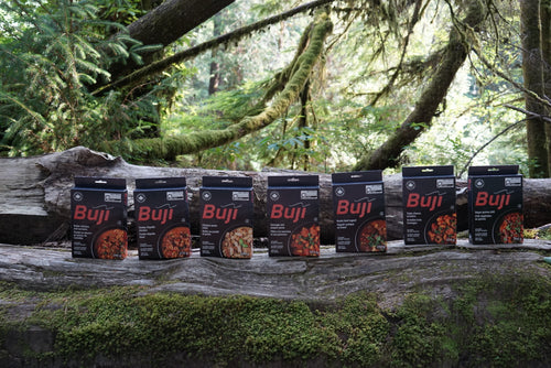 Six packages of Buji meal replacement powder arranged in a row on a mossy log in a forest setting.